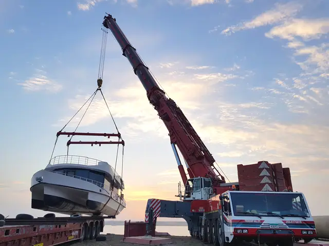 Mobile crane lifting a yacht onto a lowbed trailer at sunset